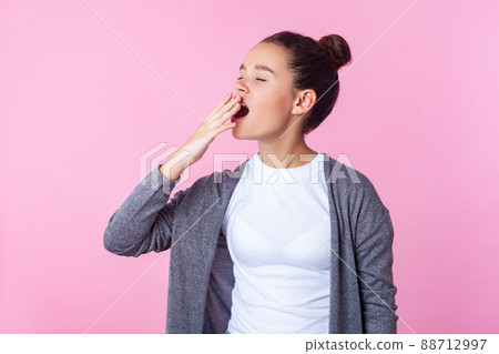 Portrait of sleepy brunette teen girl with bun hairstyle in casual clothes yawning and covering mouth with hand, feeling bored fatigued, lack of energy. indoor studio shot isolated on pink background 88712997