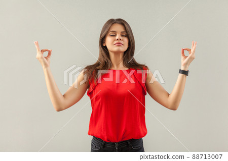 Yoga and relaxation. Portrait of calm beautiful brunette young woman in red shirt standing in yoga pose with closed eyes and meditating. indoor, studio shot, isolated on gray background. Yoga and relaxation. Portrait of calm beautiful brunette young woman in red shirt standing in yoga pose with closed eyes and meditating. indoor, studio shot, isolated on gray background. 88713007