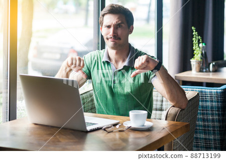 Dislike. Young dissatisfied businessman in green t-shirt sitting, working on laptop, looking at camera and showing thumbs down. business and freelancing concept. indoor shot near big window at daytime 88713199
