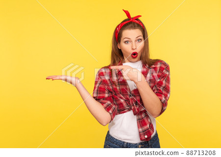 Crazy product advertising. Portrait of amazed pretty pinup girl in checkered shirt and headband pointing at copy space on her palm, empty place for commercial image. retro 50's style, studio shot 88713208