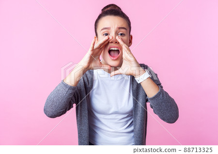 Attention. Portrait of brunette teenage girl with bun hairstyle in casual clothes holding hands near wide open mouth, loudly announcing ad, looking worried frowning. studio shot, pink background 88713325