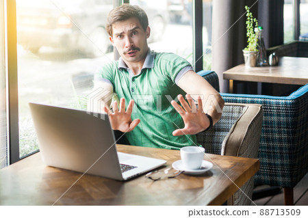 No, stop, please don't touch me. Young scared businessman in green t-shirt sitting with laptop, looking with afraid face and stop sign. business problem concept. indoor shot near big window at daytime No, stop, please don't touch me. Young scared businessman in green t-shirt sitting with laptop, looking with afraid face and stop sign. business problem concept. indoor shot near big window at daytime 88713509