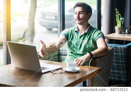 Handshake. Young satisfied businessman in green t-shirt sitting, looking at laptop screen, giving hand to greeting on interview video call meeting. business concept. indoor shot near window at daytime 88713521