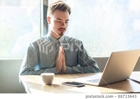 Portrait of calm handsome successful bearded young freelancer in blue shirt are sitting in cafe and having a rest, holding hands like have meditative to have emotional pleasure. indoor 88713643