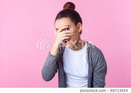 Portrait of adorable curious brunette teen girl with bun hairstyle in casual clothes peeking through fingers, looking embarrassed, pleasantly surprised. indoor studio shot isolated on pink background Portrait of adorable curious brunette teen girl with bun hairstyle in casual clothes peeking through fingers, looking embarrassed, pleasantly surprised. indoor studio shot isolated on pink background 88713654