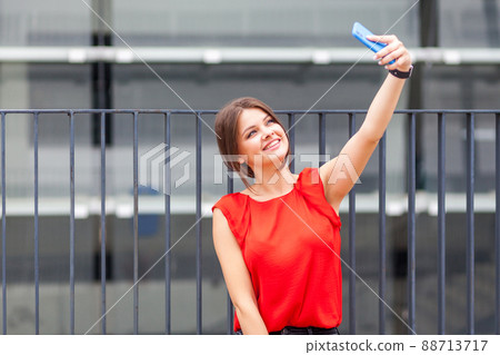 Portrait of lovely cheerful woman with brunette hair in stylish red shirt holding smartphone and taking selfie with toothy smile or blogger making live video in city outdoor, youth lifestyle Portrait of lovely cheerful woman with brunette hair in stylish red shirt holding smartphone and taking selfie with toothy smile or blogger making live video in city outdoor, youth lifestyle 88713717