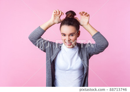 Portrait of adorable funny brunette teen girl with bun hairstyle in casual clothes showing bunny ears, laughing and having fun, childish carefree mood. indoor studio shot isolated on pink background Portrait of adorable funny brunette teen girl with bun hairstyle in casual clothes showing bunny ears, laughing and having fun, childish carefree mood. indoor studio shot isolated on pink background 88713746