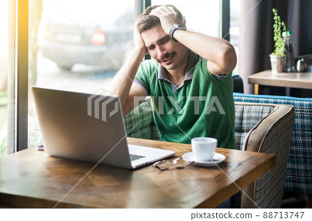 Mistake. Young sad businessman in green t-shirt sitting and working on laptop and holding his head because he made mistake. business and freelancing concept. indoor shot near big window at daytime. Mistake. Young sad businessman in green t-shirt sitting and working on laptop and holding his head because he made mistake. business and freelancing concept. indoor shot near big window at daytime. 88713747