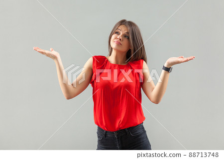 Portrait of puzzled beautiful brunette young woman in red shirt standing with raised arms, looking away thinking and confused. indoor, studio shot, isolated on gray background. 88713748