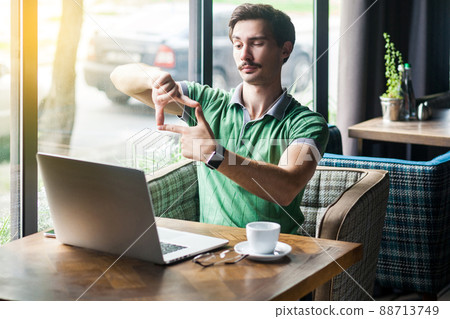 Focus on goals work. Young serious businessman in green t-shirt sitting and looking at laptop screen with crop composition gesture. business goals concept. indoor shot near big window at daytime. Focus on goals work. Young serious businessman in green t-shirt sitting and looking at laptop screen with crop composition gesture. business goals concept. indoor shot near big window at daytime. 88713749