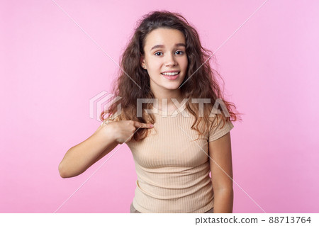 Portrait of lovely teenage girl with curly brunette hair wearing casual style beige clothes smiling at camera and pointing at herself, proud of achievements. studio shot isolated on pink background Portrait of lovely teenage girl with curly brunette hair wearing casual style beige clothes smiling at camera and pointing at herself, proud of achievements. studio shot isolated on pink background 88713764
