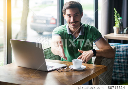 Handshake. Young happy businessman in green t-shirt sitting with laptop, toothy smile looking at camera and giving hand to greeting. business freelance concept. indoor shot near big window at daytime. 88713814