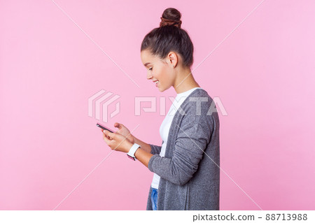 Side view of happy beautiful teenage girl with bun hairstyle in casual clothes using cell phone, feeling excited of chatting with friends, scrolling social network. indoor studio shot, pink background 88713988