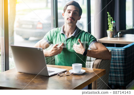 This is me. Young proud haughty businessman in green t-shirt sitting with laptop and looking at camera with proud confident face. business and freelancing concept. indoor shot near window at daytime. This is me. Young proud haughty businessman in green t-shirt sitting with laptop and looking at camera with proud confident face. business and freelancing concept. indoor shot near window at daytime. 88714143