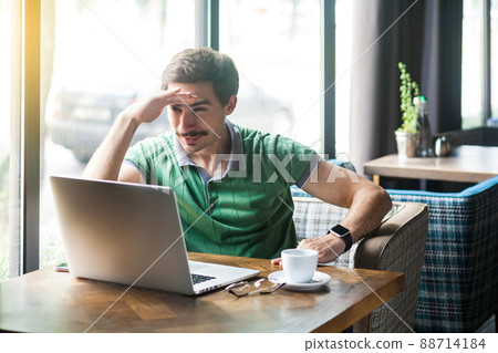 Young attentive businessman in green t-shirt sitting and working on laptop, holding hand on his forehead and looking to something. business and freelancing concept. indoor shot near window at daytime. 88714184