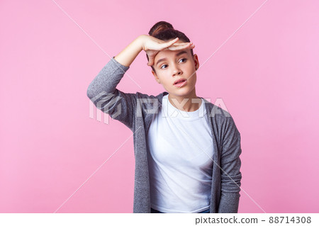 Portrait of curious brunette teenage girl with bun hairstyle in casual clothes looking into distance, holding palm over her eyes, planning far future. indoor studio shot isolated on pink background Portrait of curious brunette teenage girl with bun hairstyle in casual clothes looking into distance, holding palm over her eyes, planning far future. indoor studio shot isolated on pink background 88714308