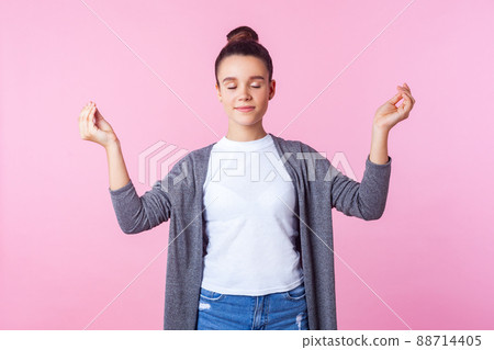 Yoga practice for mind balance. Portrait of calm brunette teenage girl with bun hairstyle in casual clothes standing with closed eyes. doing mudra gesture with fingers, meditation. indoor studio shot Yoga practice for mind balance. Portrait of calm brunette teenage girl with bun hairstyle in casual clothes standing with closed eyes. doing mudra gesture with fingers, meditation. indoor studio shot 88714405