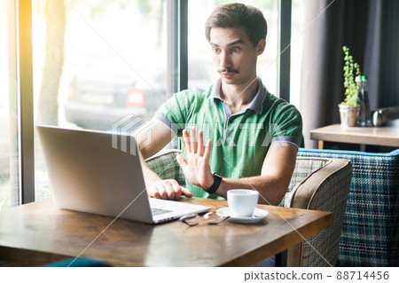 Stop it. Young serious businessman in green t-shirt sitting, working and looking at laptop screen and showing stop sign gesture. business and freelancing concept. indoor shot near window at daytime. Stop it. Young serious businessman in green t-shirt sitting, working and looking at laptop screen and showing stop sign gesture. business and freelancing concept. indoor shot near window at daytime. 88714456