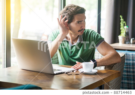 Young businessman in green t-shirt sitting and try to listen something on laptop or video call. business and freelancing concept. indoor shot near big window at daytime. Young businessman in green t-shirt sitting and try to listen something on laptop or video call. business and freelancing concept. indoor shot near big window at daytime. 88714463