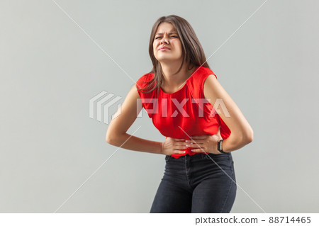 Stomach, belly pain. Portrait of sick sad brunette young woman in red shirt standing and holding her painful problem belly. indoor, studio shot, isolated on gray background. Stomach, belly pain. Portrait of sick sad brunette young woman in red shirt standing and holding her painful problem belly. indoor, studio shot, isolated on gray background. 88714465