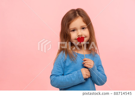 Portrait of lovely cute brunette little girl covering mouth with red paper lips or heart shape, kindly looking at camera, fashion and beauty concept. indoor studio shot isolated on pink background Portrait of lovely cute brunette little girl covering mouth with red paper lips or heart shape, kindly looking at camera, fashion and beauty concept. indoor studio shot isolated on pink background 88714545