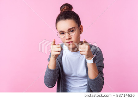 Hey you. Portrait of sophisticated brunette teenage girl with bun hairstyle in round eyeglasses pointing at camera with serious smart expression. indoor studio shot isolated on pink background Hey you. Portrait of sophisticated brunette teenage girl with bun hairstyle in round eyeglasses pointing at camera with serious smart expression. indoor studio shot isolated on pink background 88714626
