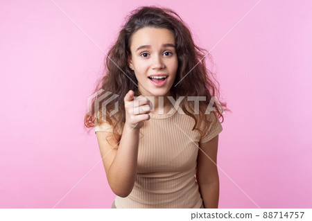 Hey you. Portrait of surprised teen girl with curly brunette hair wearing casual style beige clothes pointing at camera, standing with open mouth, wondered astonished expression. studio shot, indoor 88714757