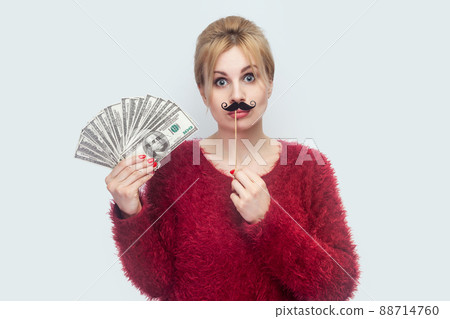 Beautiful young woman in red blouse standing, demonstrate fan of money, holding manlike mustache sticker, looking at camera with big eyes. Indoor, studio shot, isolated on gray background Beautiful young woman in red blouse standing, demonstrate fan of money, holding manlike mustache sticker, looking at camera with big eyes. Indoor, studio shot, isolated on gray background 88714760