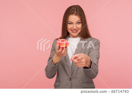 Portrait of positive woman employee in suit jacket holding sweet doughnut and pointing to camera, showing unhealthy snack, junk food for business people. indoor studio shot isolated on pink background 88714793
