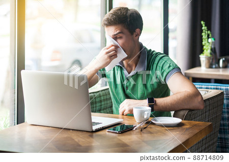 Young cold sick businessman in green t-shirt sitting at work, sneezing and cleaning his nose with tissue napkin. business and helthcare concept. indoor shot near big window at daytime. 88714809