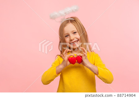 Portrait of sweet lovely angelic little girl with halo over head holding toy hearts and looking at camera with toothy smile, symbol of love and charity. indoor studio shot isolated on pink background 88714904