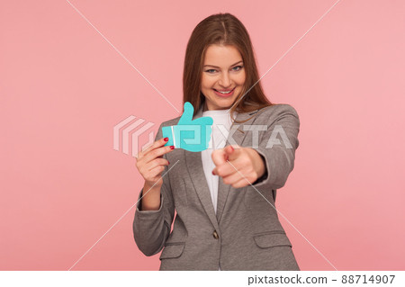 Portrait of cheerful businesswoman in suit jacket pointing to camera and holding blue thumbs up icon, gesturing hey you, comment and push like button, support blog. indoor studio shot, pink background 88714907