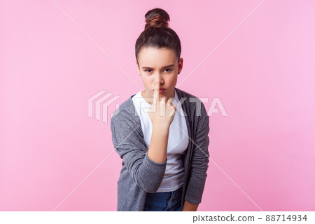 You are liar. Portrait of cute brunette teenage girl with bun hairstyle in casual clothes touching nose with finger showing lie gesture, expressing disbelief, suspicion. studio shot, pink background 88714934
