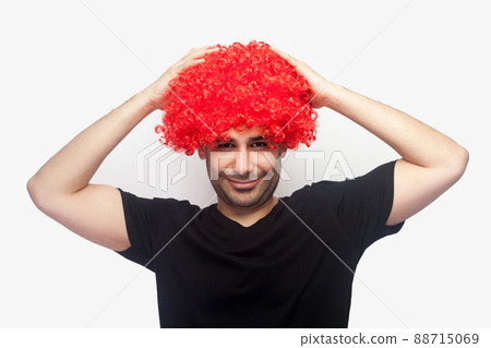 Portrait of crazy hipster, joyful man with bristle and in t-shirt touching red wig on his head and smiling at camera, looking funny with bright hair. indoor studio shot isolated on white background Portrait of crazy hipster, joyful man with bristle and in t-shirt touching red wig on his head and smiling at camera, looking funny with bright hair. indoor studio shot isolated on white background 88715069
