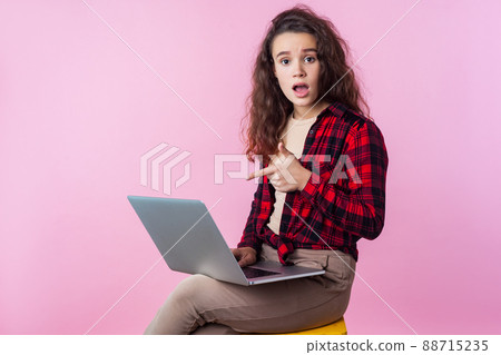 Portrait of frustrated computer user, teen girl with curly brunette hair in plaid shirt pointing at laptop, looking worried about software error, program bug. studio shot isolated on pink background Portrait of frustrated computer user, teen girl with curly brunette hair in plaid shirt pointing at laptop, looking worried about software error, program bug. studio shot isolated on pink background 88715235
