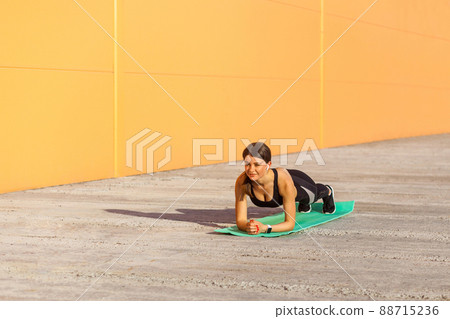 Muscular sporty strong beautiful young athletic woman in black sportwear standing on perfect plank position on elbows on green mat. Outdoor, orange wall background, sport and healthy concept 88715236