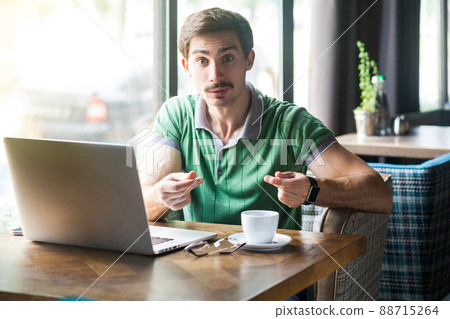 Give me money. Young businessman in green t-shirt sitting and looking at camera with money gesture and want his salary. business and freelancing concept. indoor shot near big window at daytime. 88715264