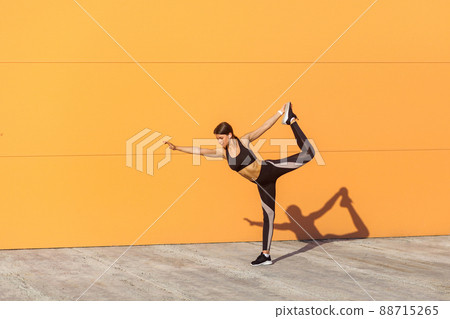 Young attractive girl practicing yoga, standing in natarajasana exercise, holding balance, working out, wearing black sportswear, urban style, full length, orange background, outdoor sport concept 88715265
