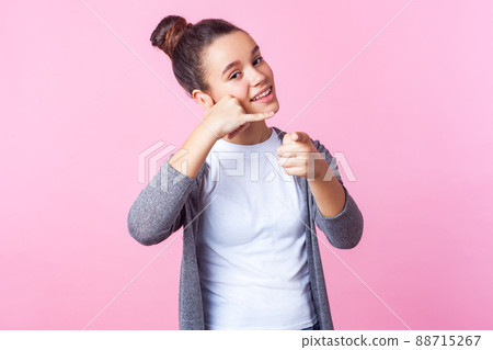 Hey you, call me. Portrait of adorable brunette teen girl with bun hairstyle in casual clothes smiling playfully, making telephone gesture and pointing at you. studio shot isolated on pink background 88715267