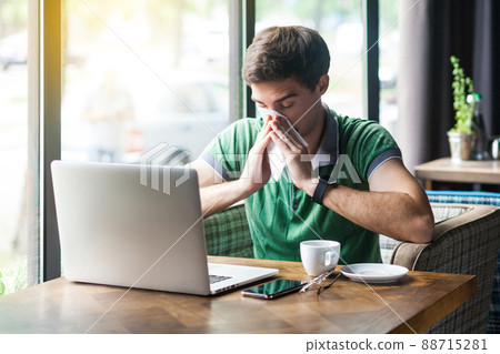 Young cold sick businessman in green t-shirt sitting at work, sneezing and cleaning his nose with tissue napkin. business and helthcare concept. indoor shot near big window at daytime. 88715281