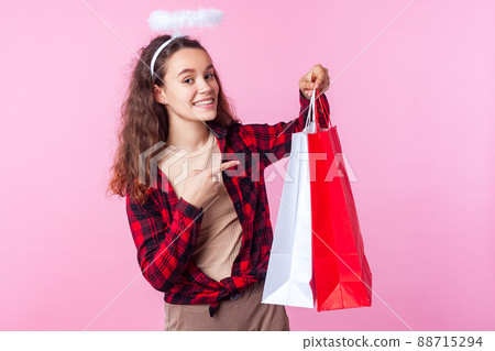 Look at my shopping bags. Portrait of angelic teenage girl with curly brunette hair and toy halo above head pointing at packages boasting purchase, holiday sales. studio shot isolated, pink background Look at my shopping bags. Portrait of angelic teenage girl with curly brunette hair and toy halo above head pointing at packages boasting purchase, holiday sales. studio shot isolated, pink background 88715294
