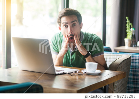 It's unbelievable. Young shocked businessman in green t-shirt sitting and working on laptop, looking at camera with surprised face. business and freelancing concept. indoor shot near window at daytime It's unbelievable. Young shocked businessman in green t-shirt sitting and working on laptop, looking at camera with surprised face. business and freelancing concept. indoor shot near window at daytime 88715295