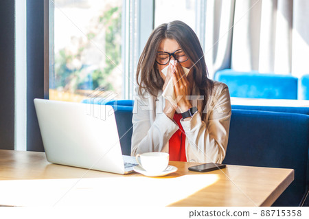 Sneeze, cold, influenza or allergies. Portrait of sick stylish brunette young woman in glasses sitting holding tissue napkin and cleaning her nose. indoor studio shot, cafe, office background. Sneeze, cold, influenza or allergies. Portrait of sick stylish brunette young woman in glasses sitting holding tissue napkin and cleaning her nose. indoor studio shot, cafe, office background. 88715358