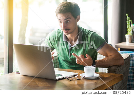 Young angry crazy businessman in green t-shirt sitting, working, looking at laptop screen and showing middle finger fuck sign. business and freelancing concept. indoor shot near big window at daytime. 88715485