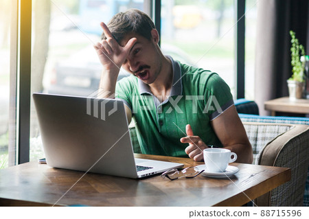 I am loser. Young angy businessman in green t-shirt sitting and working on laptop with loser gesture on his forehead and screaming. business and freelancing concept. indoor shot near window at daytime I am loser. Young angy businessman in green t-shirt sitting and working on laptop with loser gesture on his forehead and screaming. business and freelancing concept. indoor shot near window at daytime 88715596