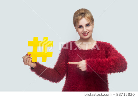 Portrait of happy young woman in red blouse standing and holding large big yellow hashtag sign and pointing finger, social media concept. Indoor, isolated, studio shot, copy space, gray background 88715820