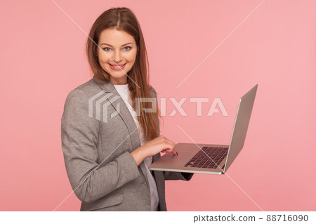 Portrait of elegant positive businesswoman in suit jacket holding laptop and smiling to camera, using computer to study, working online, doing job remotely. studio shot isolated on pink background Portrait of elegant positive businesswoman in suit jacket holding laptop and smiling to camera, using computer to study, working online, doing job remotely. studio shot isolated on pink background 88716090