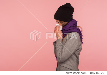 Seasonal influenza. Side view of flu-sick woman in warm hat and scarf blowing nose in tissue, sneezing suffering allergy symptoms, feeling unhealthy. indoor studio shot isolated on pink background 88716122