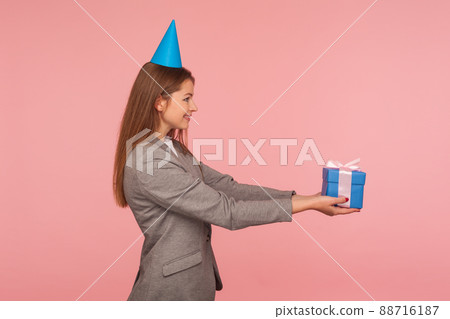 Happy birthday, take present. Side view of happy woman in business suit and with party cone on her head giving gift box, congratulating on birthday anniversary, job promotion. studio shot, isolated Happy birthday, take present. Side view of happy woman in business suit and with party cone on her head giving gift box, congratulating on birthday anniversary, job promotion. studio shot, isolated 88716187