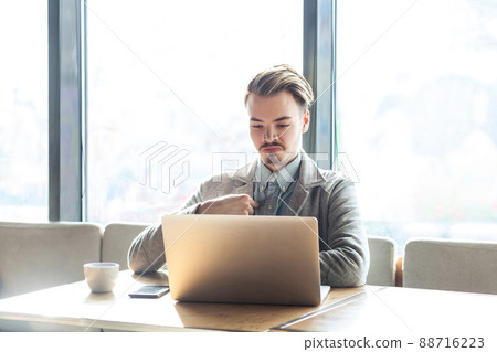 Portrait of confident selfish satisfied egoistic bearded young freelancer in grey blazer are sitting in cafe, working on laptop and pointing finger at himself with pride. indoor, inside Portrait of confident selfish satisfied egoistic bearded young freelancer in grey blazer are sitting in cafe, working on laptop and pointing finger at himself with pride. indoor, inside 88716223